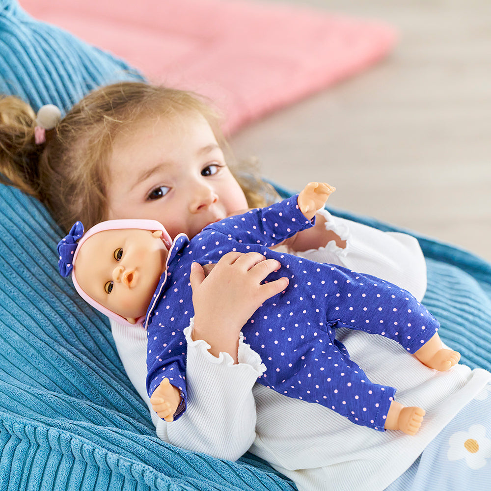 Une enfant câline son poupon Bébé Calin Capucine en robe bleue à pois sur un canapé.