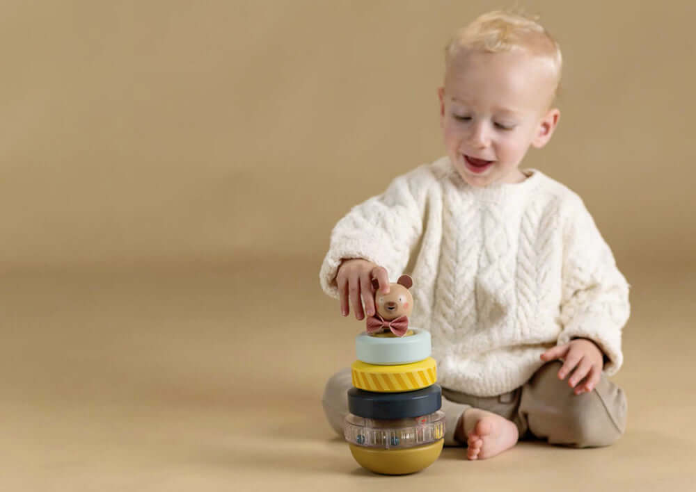 Un enfant joyeux empile les anneaux à empiler Taf Toys avec un ourson coloré.