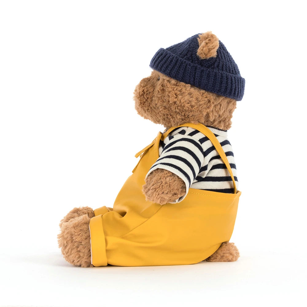 Jellycat Bartholomew Bear outfit avec un chapeau bleu marine et une combinaison jaune, assis sur un fond blanc.