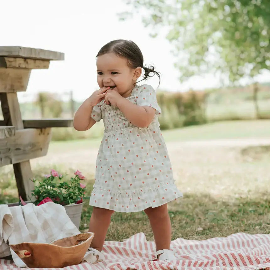 bébé souriant en robe smockée bébé Vichy fraises dans un jardin, profitant d'une journée ensoleillée.